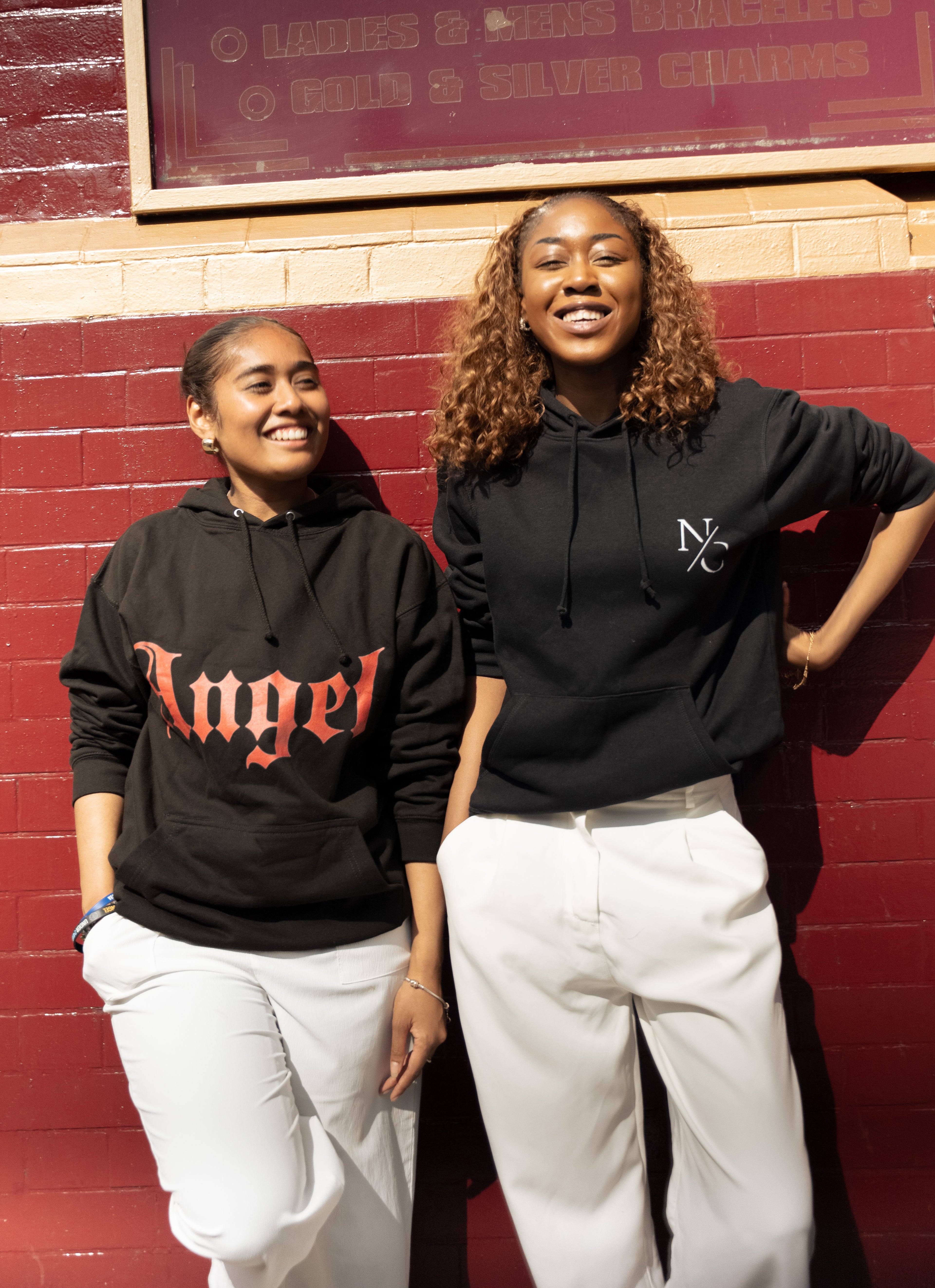Two individuals wearing black hoodies with visible text standing against a red brick wall.